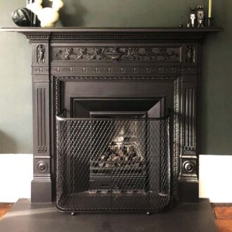 A black metal wirework fireguard stands in front of a traditional open fireplace.